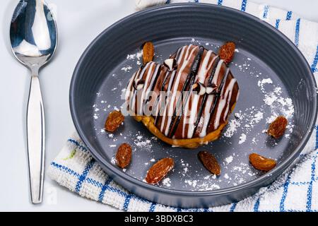 Chocolate choux cream pastry with raisins in black ceramic dish on kitchent cloth and metal spoon on white background. Stock Photo