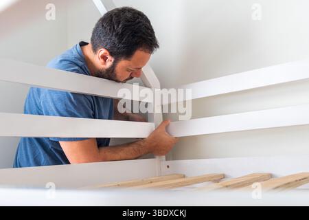 A man is fixing a bed frame. The bed is white and the frame is made of wood Stock Photo