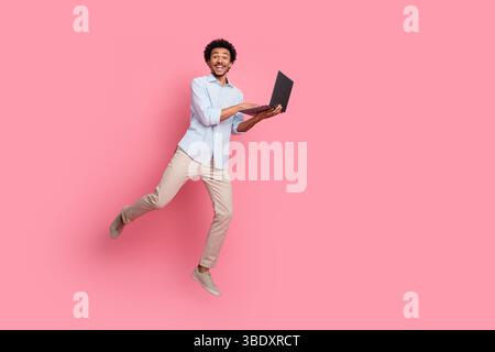 Excited young man in casual attire jumping while using a laptop on pink background showcasing energy and enthusiasm Stock Photo