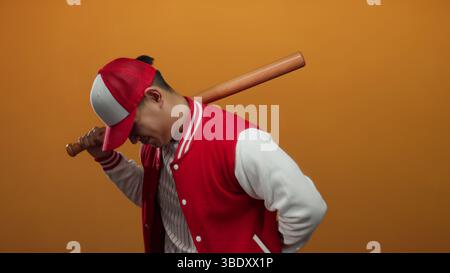 Young man in a baseball uniform poses confidently with a bat against an orange background, showcasing a sporty and vibrant atmosphere. Stock Photo