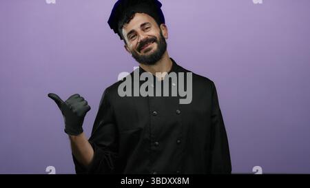 Hispanic bearded chef in black attire smiles against a vibrant purple backdrop, conveying a sense of culinary pride and inviting warmth with a thumbs- Stock Photo