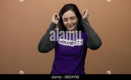 Woman dancing in purple volunteer shirt against orange background smiles joyfully, showcasing volunteerism, happiness, and positivity, with hands rais Stock Photo