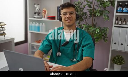 Young man in medical scrubs with headset smiles confidently while using laptop in hospital clinic office with organized shelves highlighting a profess Stock Photo