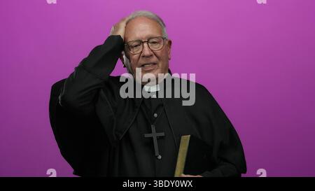 Senior priest in black robe, holding a book, stands over an isolated pink background with a thoughtful expression, highlighting faith and contemplatio Stock Photo