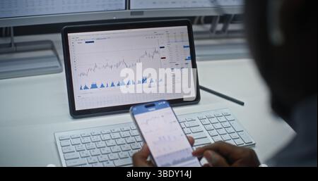 African American Stock Exchange Trader Analyzing Real-Time Stocks and Exchange Market Charts on Smartphone and Tablet Computer Using Online Trading App. Investment Agent Working in Office. Close Up. Stock Photo