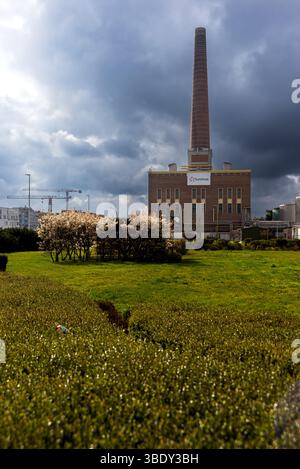 30.03.2025 Gent, Belgium, a tall brick smokestack rises from a Luminus power station under dramatic cloudy skies with green lawns, trimmed bushes, and Stock Photo