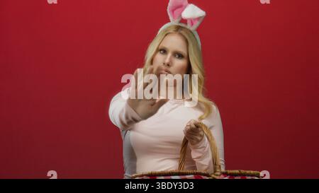 Young blonde woman with bunny ears and pink shirt makes stop gesture against red background, holding picnic basket humorously. Stock Photo