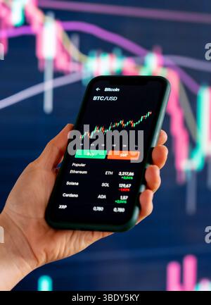 Smartphone displaying crypto trading app with financial charts in background Stock Photo