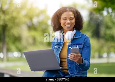 Happy Black student woman using smartphone and laptop outdoors Stock Photo