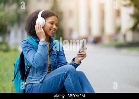 Black Female Student Listening To Music While Sitting On Grass Outdoors Stock Photo