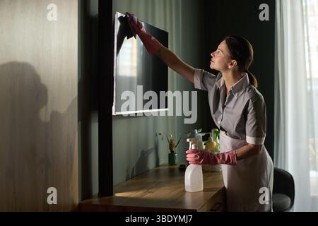 Female professional wearing grey uniform and pink gloves cleaning television with spray bottle on wooden cabinet in dimly lit room Stock Photo