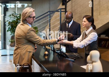 Businessmen interacting with hotel receptionist, engaging in check-in process. One businessman in tan suit handing documents, receptionist confirming details, other businessman reviewing forms Stock Photo
