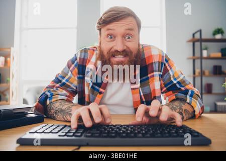 Focused young man in checkered shirt types energetically on keyboard in bright cozy interior Stock Photo