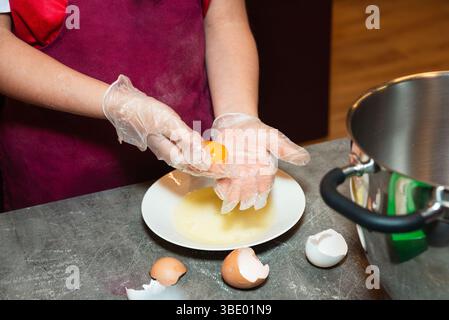 A person wearing gloves separates a raw egg yolk from the white over a plate, preparing ingredients in a kitchen setting. Stock Photo