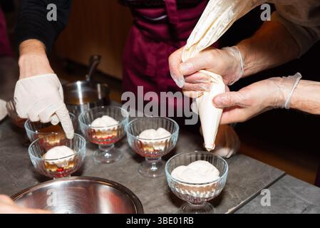 Hands in gloves using a piping bag to add whipped cream into individual glass bowls with layered dessert in a kitchen. Stock Photo