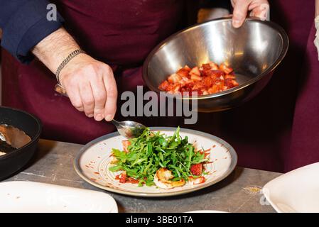 Chef preparing a gourmet salad with arugula, grilled shrimp, and strawberries, using fresh ingredients in a restaurant kitchen. Stock Photo