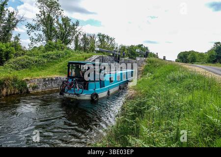 A narrowboat travels through a lock on the Royal Canal near Cloondara, County Longford, Ireland. Peaceful rural scene with lush greenery and towpath n Stock Photo
