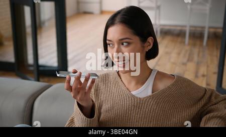Happy joyful smiling Caucasian hispanic woman girl homeowner lady female user relax on home couch record audio message smartphone voice mail talking Stock Photo