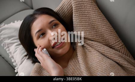 Smiling girl Caucasian hispanic latin woman female homeowner lying on couch sofa talking mobile phone answer smartphone call casual talk friendly Stock Photo