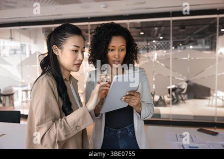 Two young multiethnic women workmates using tablet standing in workspace Stock Photo