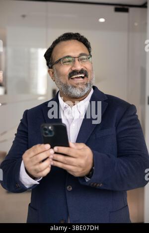 Happy Indian businessman in suit standing in workplace with smartphone Stock Photo