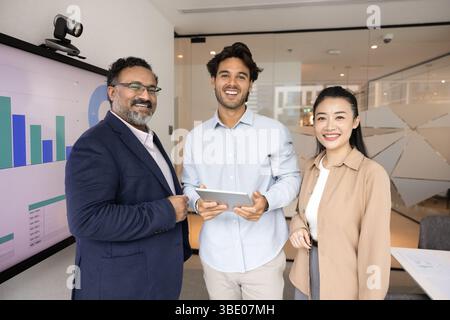 Three professionals standing in office next to screen displaying graphs Stock Photo