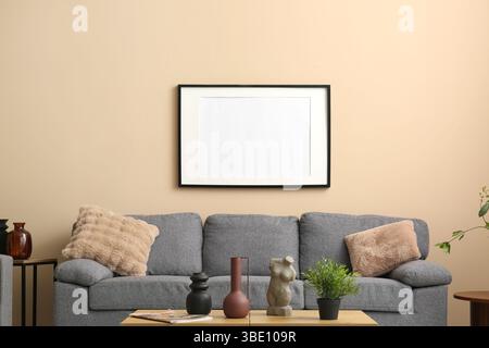 Comfortable grey sofa with soft pillows and blank photo frame hanging on beige wall in living room Stock Photo
