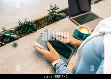 Close-up of woman holding smartphone and notebook in cafe with coffee and laptop Stock Photo
