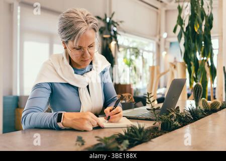 Focused senior woman with gray hair writing in notebook while working on laptop in cozy modern cafe Stock Photo