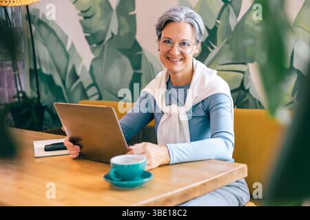 Mature woman working on freelance project in bright coffee shop, smiling and using laptop Stock Photo