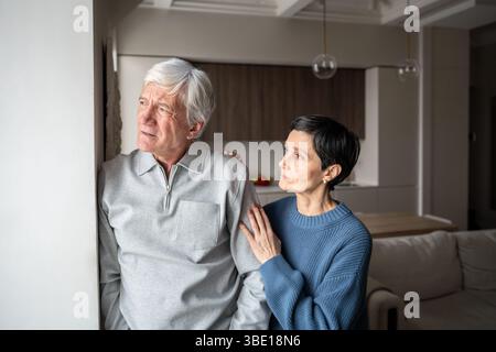 Confused senior man in thoughts with distrust look out window, wife attempts emotional connection Stock Photo