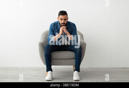 Arab man sitting in armchair with dull face expression, feeling sad or tired, having problem, suffering from depression Stock Photo