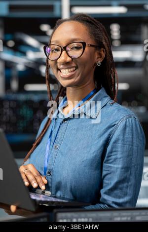 Portrait of smiling technician in data center monitoring system performance with laptop, identifying bottlenecks. Happy server hub employee troubleshooting errors, implementing optimizations Stock Photo