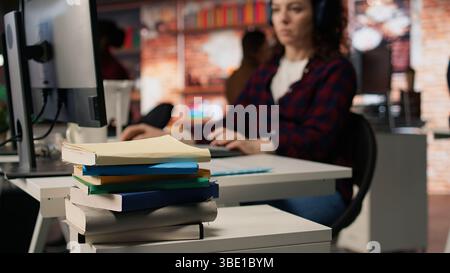 Focus on stack of books on startup office desk with woman in blurred background typing on PC, solving tasks. Pile of books in venture company workspace in front of employee working, camera A closeup Stock Photo
