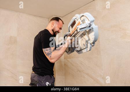 Professional tiler skillfully installing large format tiles on a wall, utilizing a tile cutter for precise cuts during the renovation process Stock Photo