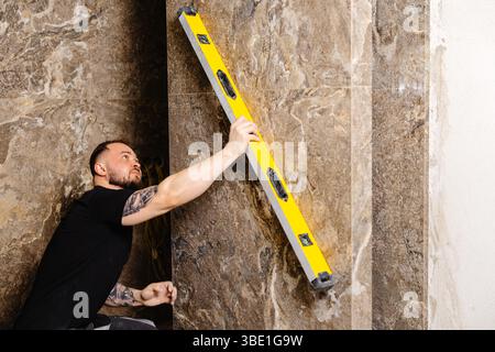 Builder checking the evenness of marble tiles with a level tool, ensuring precision and accuracy during renovation work on a wall Stock Photo