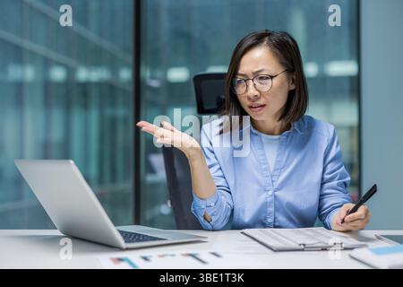 A focused asian businesswoman gestures while working on her laptop, looking puzzled, and holding a pen near a notepad. Stock Photo