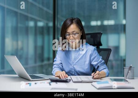A professional woman works at a desk, focused on paperwork with a laptop nearby and calculator in use. She wears glasses. Stock Photo