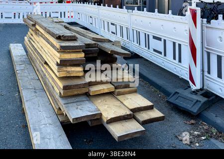 Various wooden planks are neatly stacked on a city street near a construction site, surrounded by a temporary barricade. The area appears to be underg Stock Photo