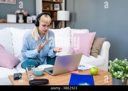 Young woman wearing headphones having video call on laptop, sitting on cozy couch at home, working remotely or studying online, gesturing while talkin Stock Photo