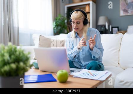 Young woman wearing headphones having video call on laptop, sitting on cozy couch at home, working remotely or studying online, gesturing while talkin Stock Photo