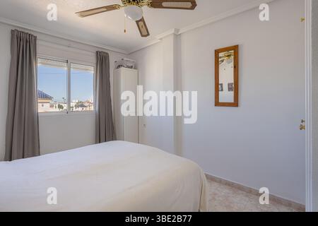 A bright master bedroom featuring a large bed, ceiling fan, window with curtains, and soft neutral decor. Stock Photo