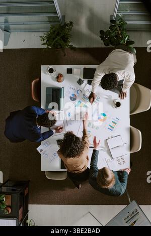 Diverse team collaborating over business data and charts on rectangular table with laptops and notebooks. Office environment fostering teamwork and br Stock Photo
