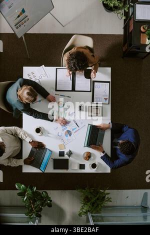 Team of diverse professionals collaborating in office, analyzing financial documents and charts on laptop screens and paper. Overhead view showing eng Stock Photo