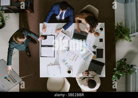 Group of professionals collaborating around table, reviewing documents and charts, discussing business strategy during meeting. Various laptops and do Stock Photo