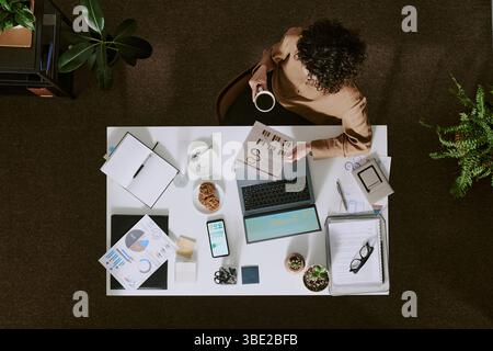 Overhead shot of office workspace with person holding coffee cup, showcasing various office supplies and tech devices including laptop, smartphone, an Stock Photo