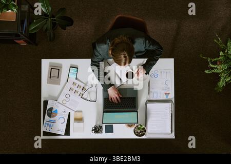 Overhead view of businessman analyzing data on reports and charts, checking details on laptop while drinking coffee at desk in office environment Stock Photo