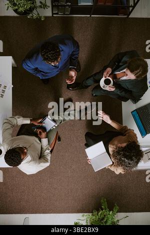 Overhead view of diverse professionals standing in office having discussion. Colleagues holding documents and coffee cups while engaging in conversati Stock Photo