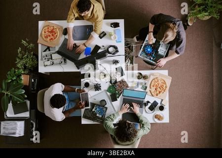 Group of colleagues engaged in collaborative meeting, working on multiple tech devices and enjoying pizza. Overhead view features diverse team with la Stock Photo