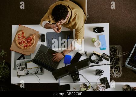 Young man sitting at desk eating pizza while working surrounded by laptops, plant, various gadgets, and drink cup. Desk appearing busy with work and e Stock Photo